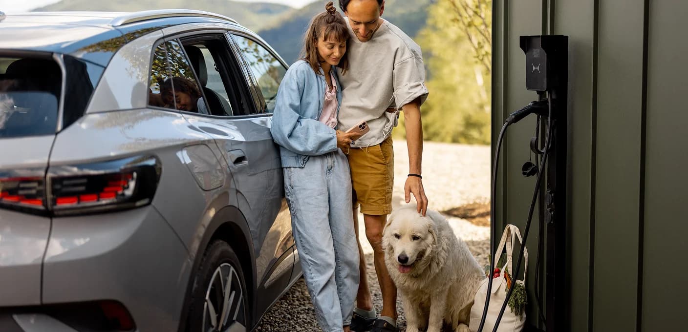 A couple beside an SUV at an EV charger, looking at a phone, with a white dog at their feet.