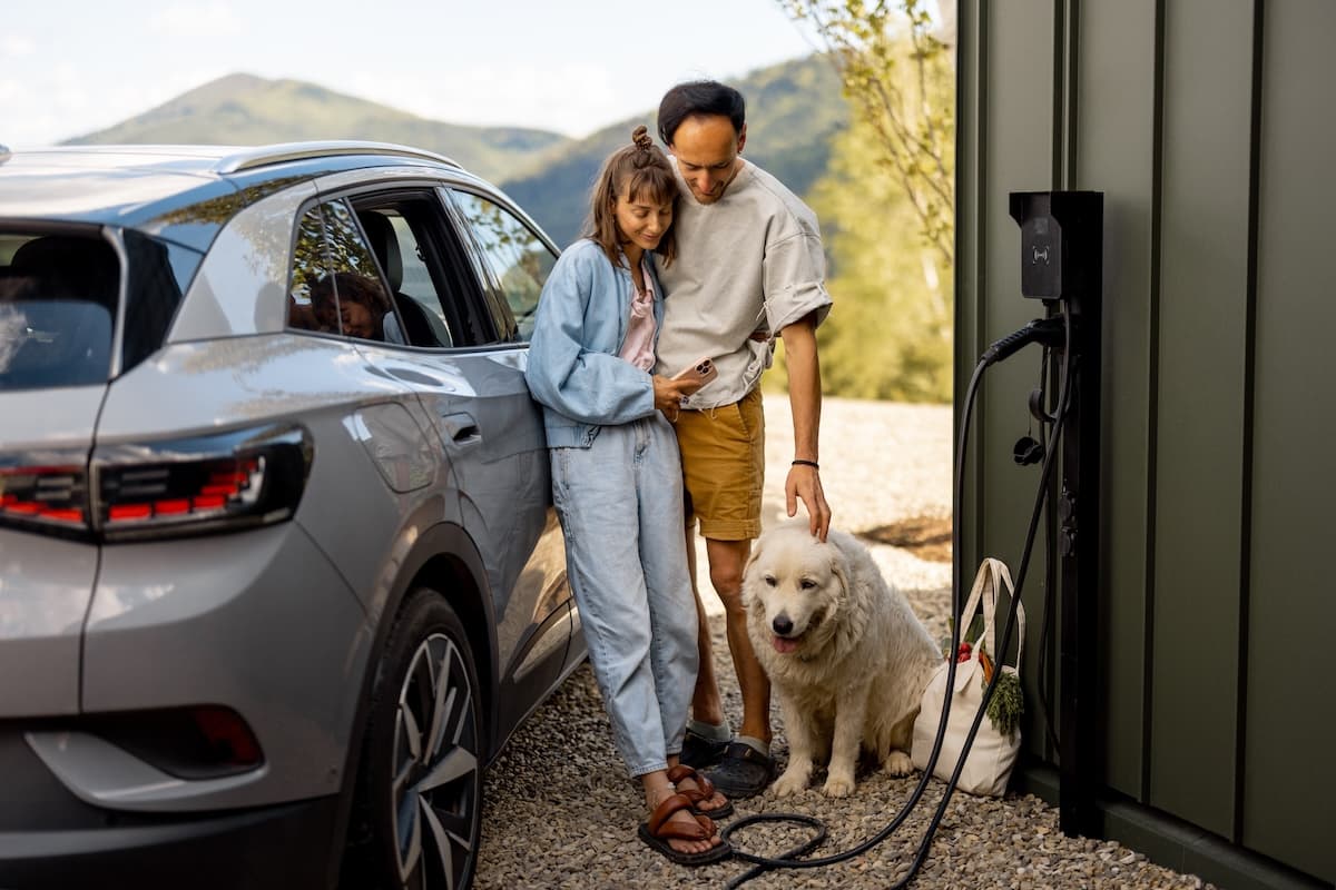 A couple beside an SUV at an EV charger, looking at a phone, with a white dog at their feet.