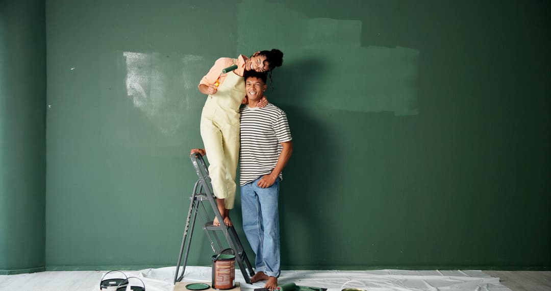 A smiling couple painting a dark green wall, one standing on a step ladder holding a paint roller.