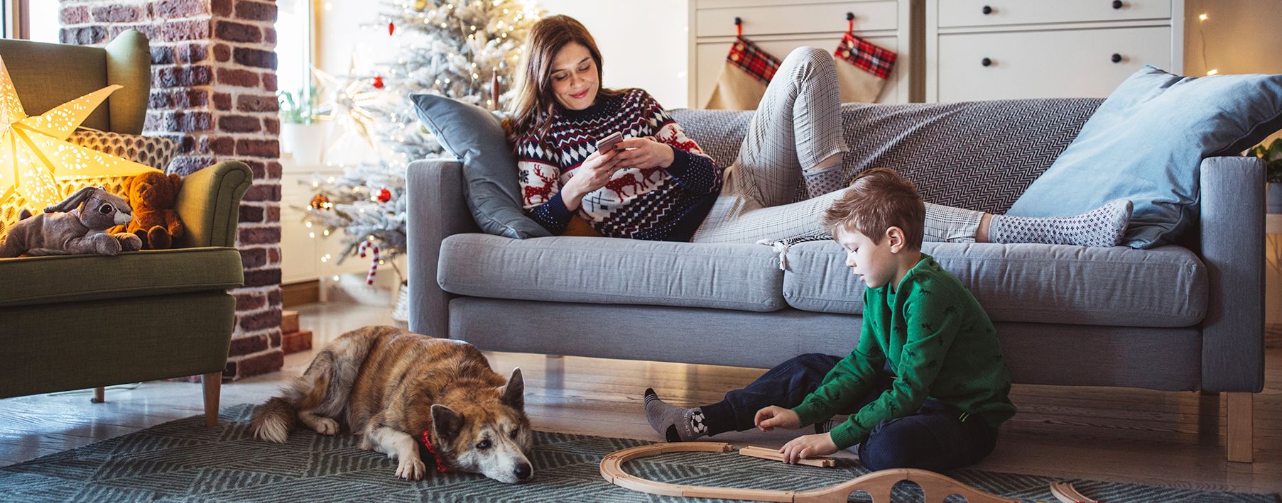 A boy playing with train tracks on a rug next to a dog, with a woman looking on her phone on a couch behind him