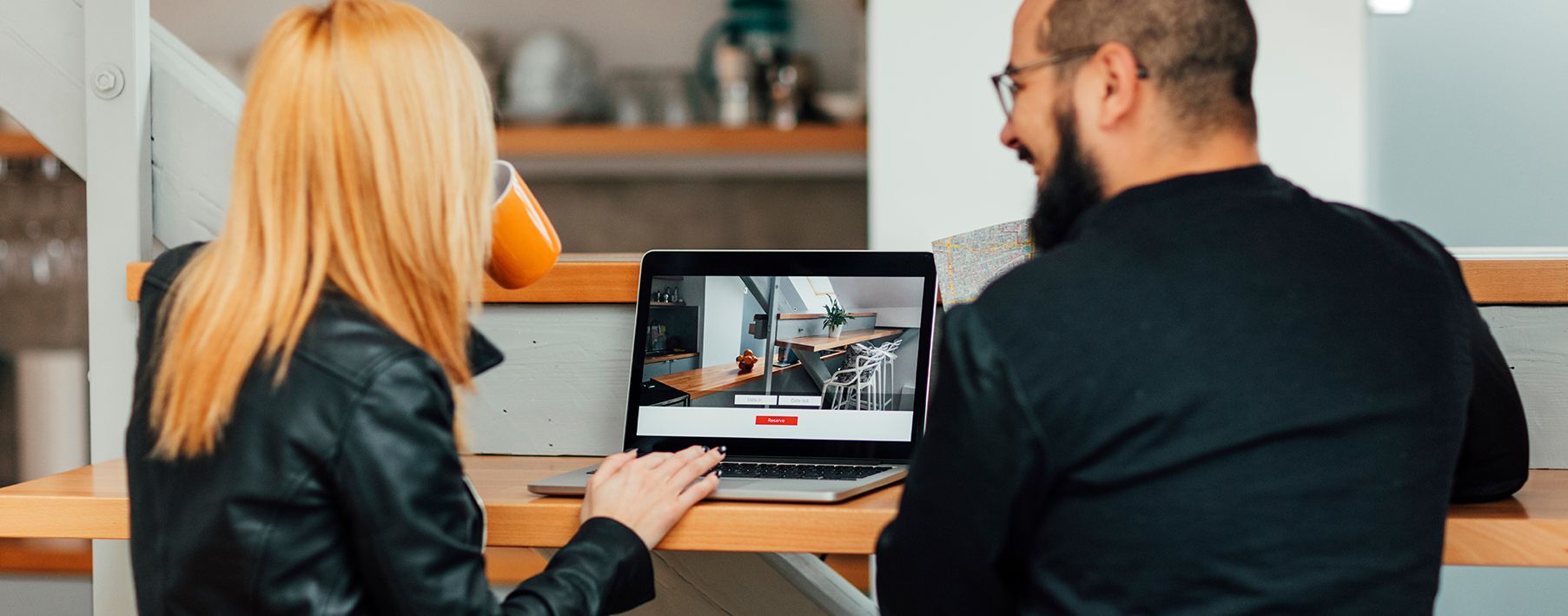 A couple sits at a desk, facing away from the camera, looking at an image of a home on the computer in front of them.