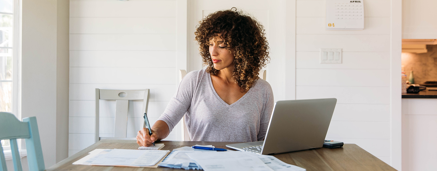 Woman sits at a desk in front of a computer; her right hand is writing on a piece of paper with a pen.