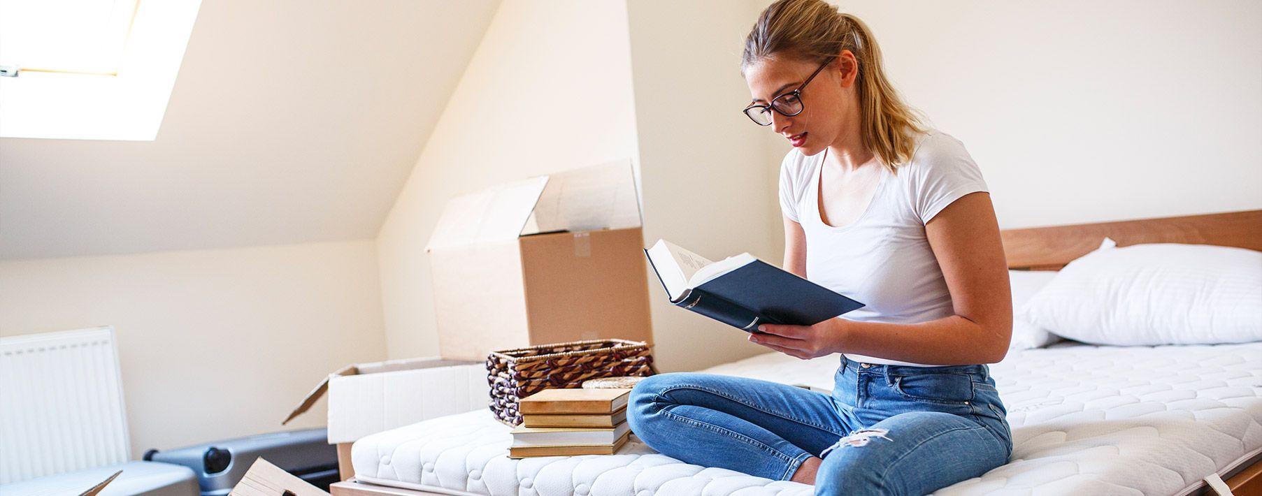 A woman sitting on a bed reading a book with boxes behind her