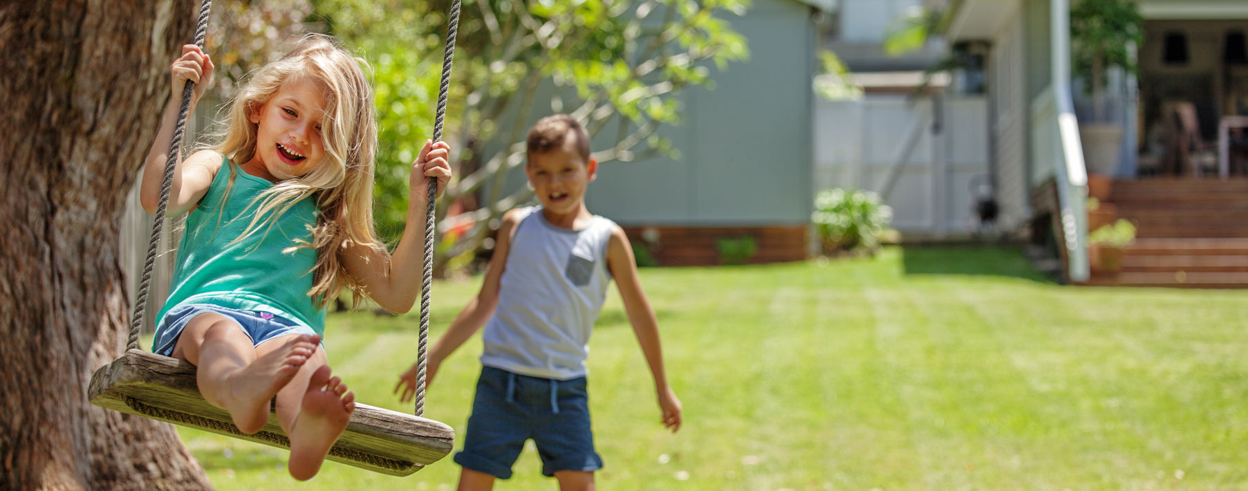 A young boy with brown hair pushes a young girl with blonde hair on a swing. Both are smiling. 