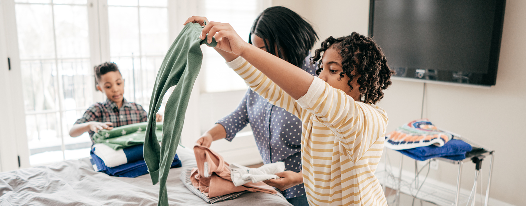 A young boy, middle-aged woman and young girl fold laundry on a bed. The young girl, closest to the camera, is holding green pants.