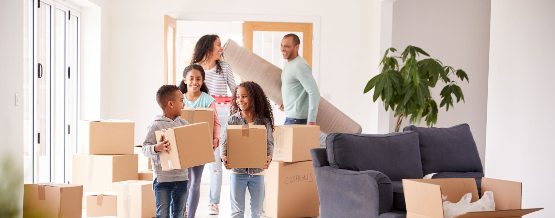 A family moves boxes into a home. A young boy and girl are in front smiling at each other, followed by another girl. The mom and dad are in the back of the room laughing. 