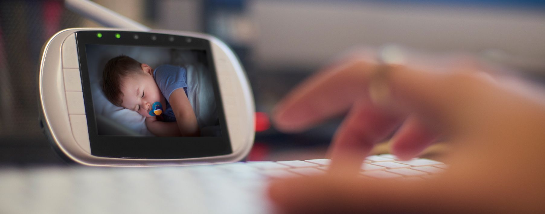 Image of a hand typing on a keyboard with a baby monitor in the background showing a sleeping baby