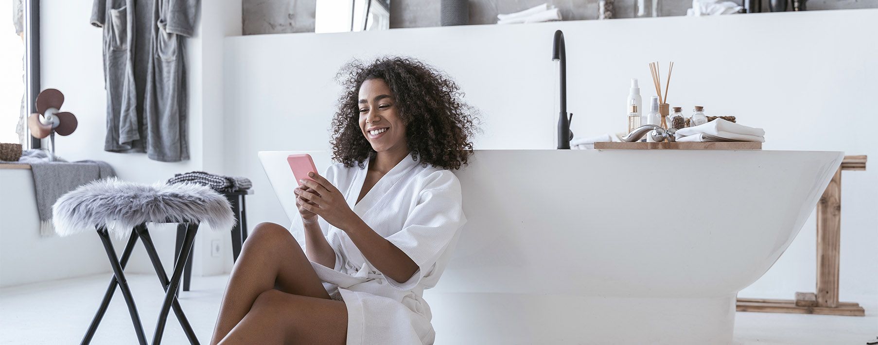 A woman sitting on the floor of her bathroom in a robe leaning on her tub while looking at her phone