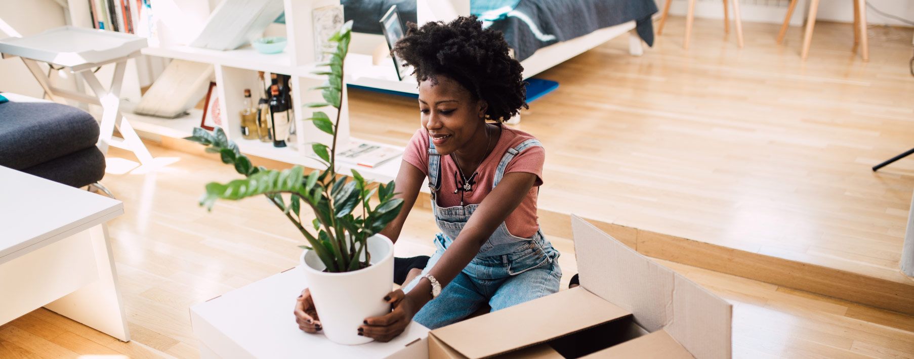 A woman in overalls sitting on the floor holding a plant with boxes nearby