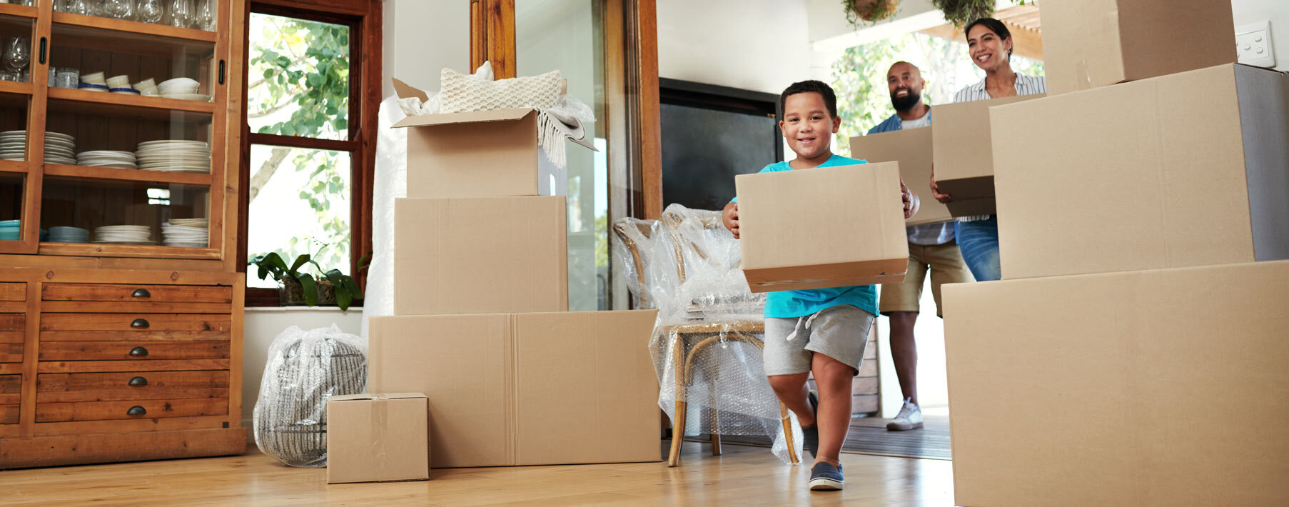 A little boy holding a moving box runs through a living room filled with moving boxes.