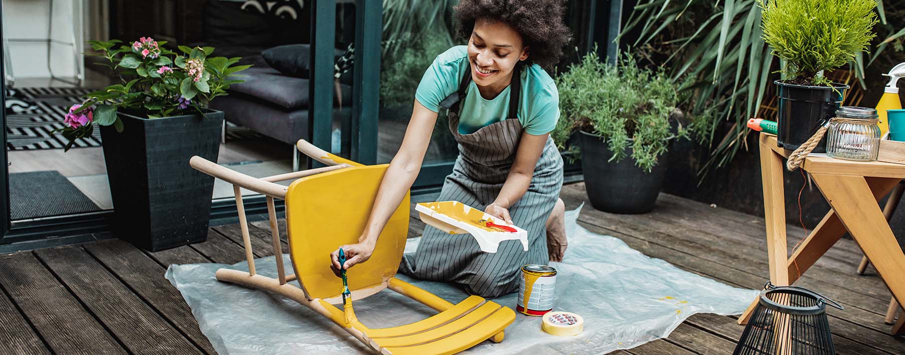 A woman paints a chair yellow on her wooden deck