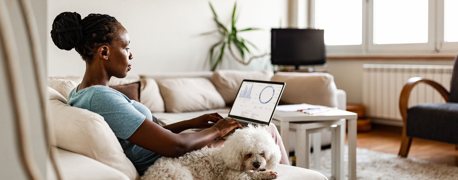 A Woman sits on a couch working on a laptop with a dog curled up by her side