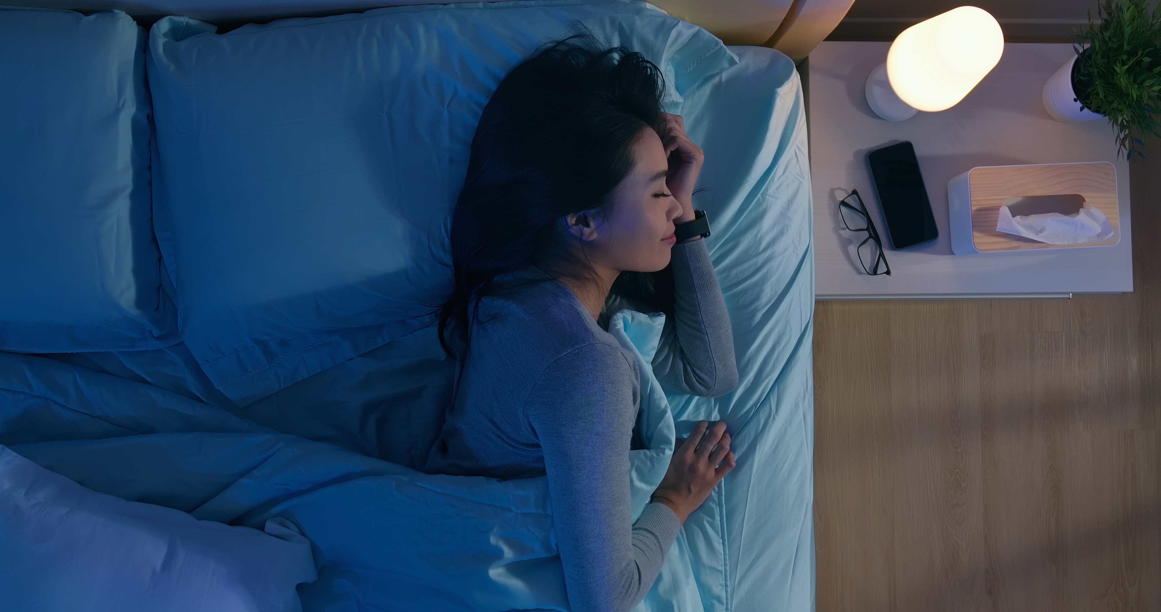 Overhead view of a woman sleeping in bed with a smartwatch on her wrist and a phone and light on the nightstand