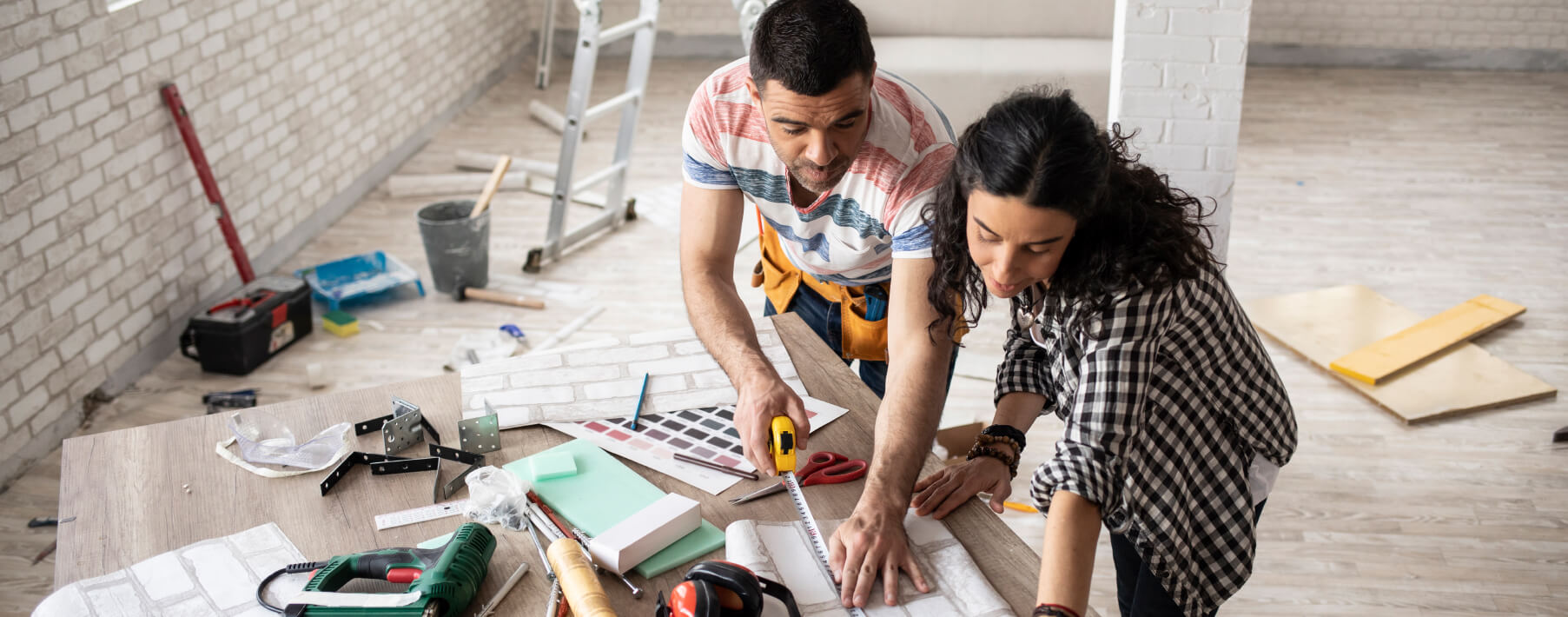 A man and a woman look over blueprint plans for a home. The man is measuring the plans with a tape measure. They are surrounded by tools. 