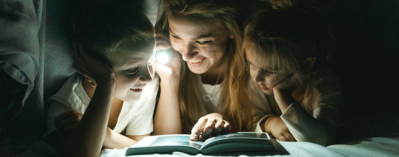 homeowner looking at books under the covers with their children while holding a flashlight