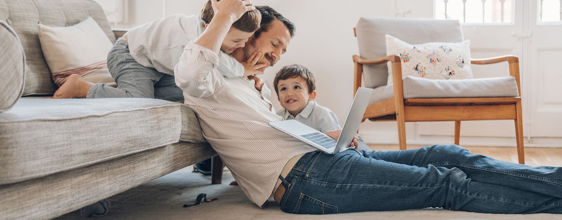 A dad sitting on a rug looking at a laptop with his sons playing around him