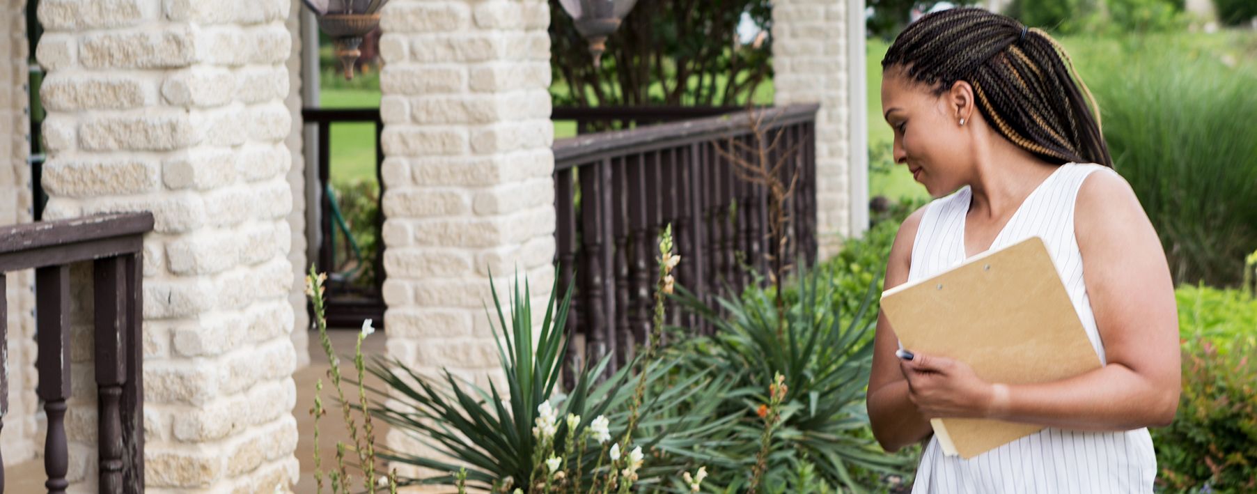 A woman holding a clipboard looking at the ground in front of a home