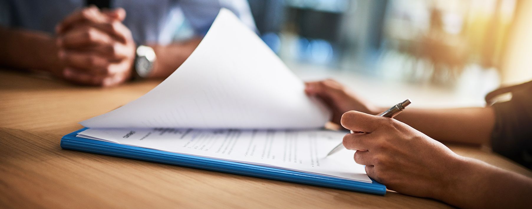 Image of a woman's hand signing a document with a mans hands in the background