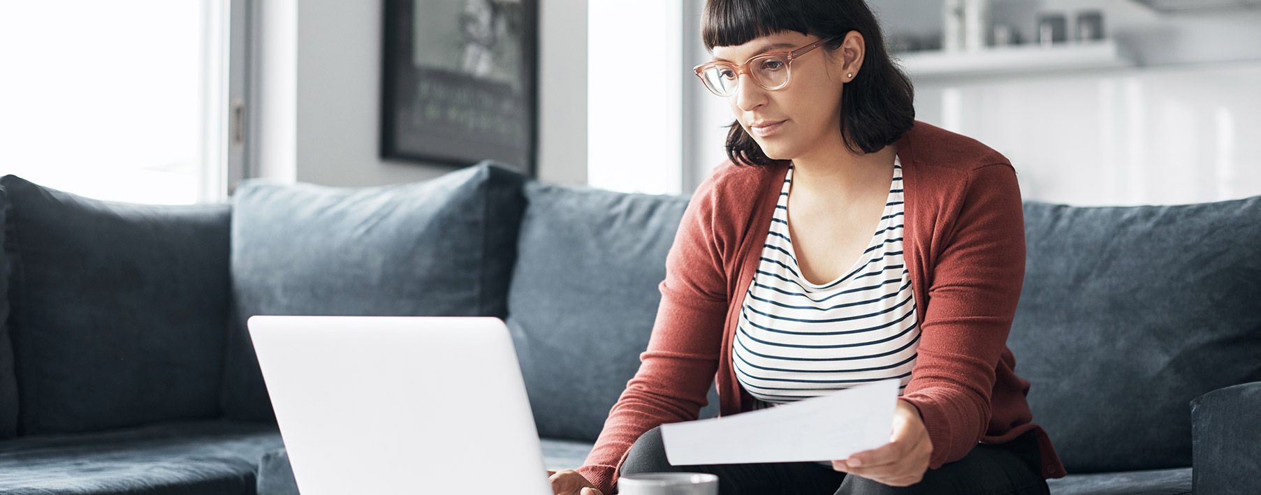 A young woman sitting on a couch looking at her laptop and a piece of paper