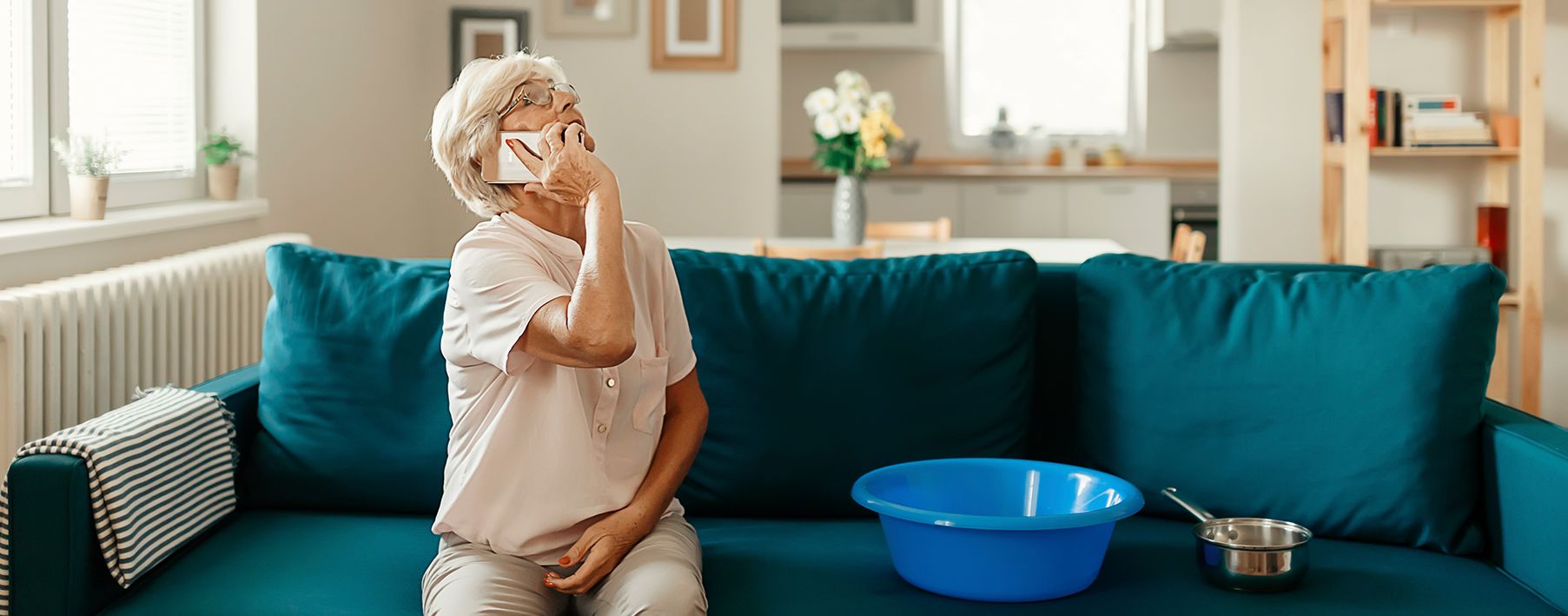 Older woman sitting on her couch on the phone, looking up at a leak in the ceiling. 