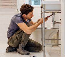 man kneeling at a sink, using a tool on the pipes