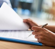 Image of a woman's hand signing a document with a mans hands in the background