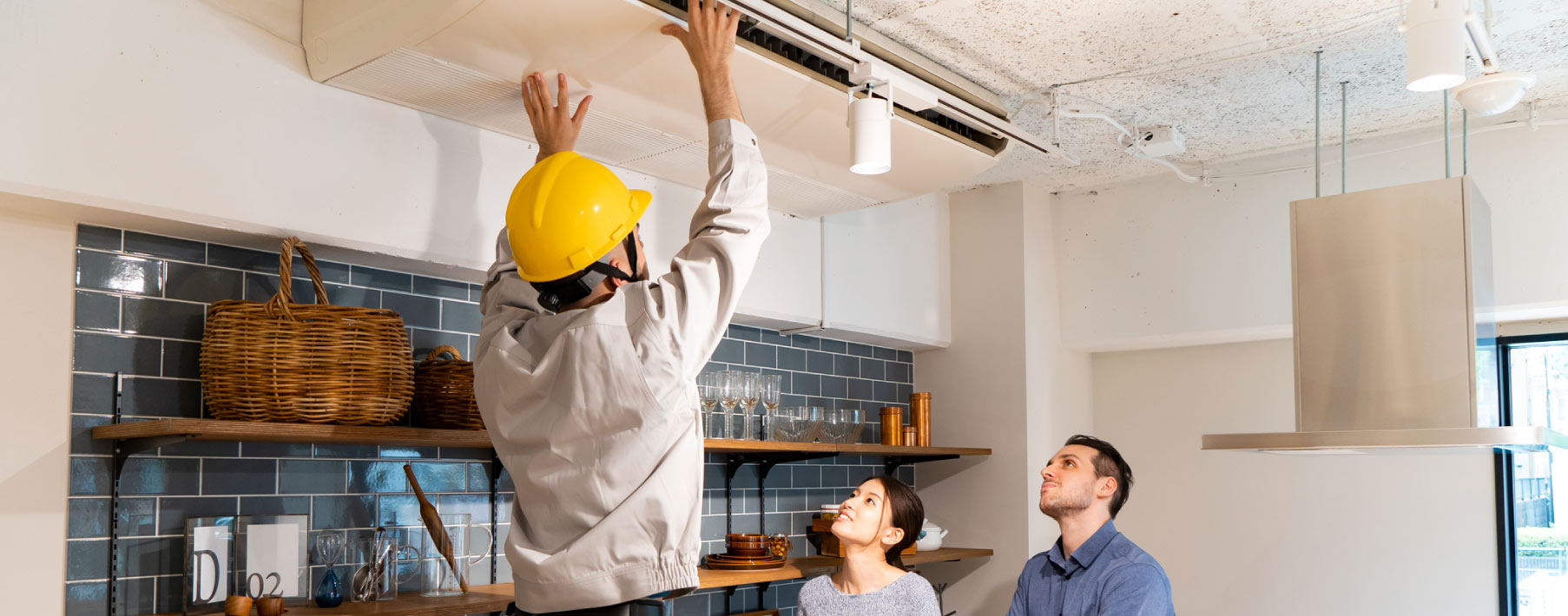 A worker with a hard hat working on something on the ceiling in the kitchen with a couple looking on
