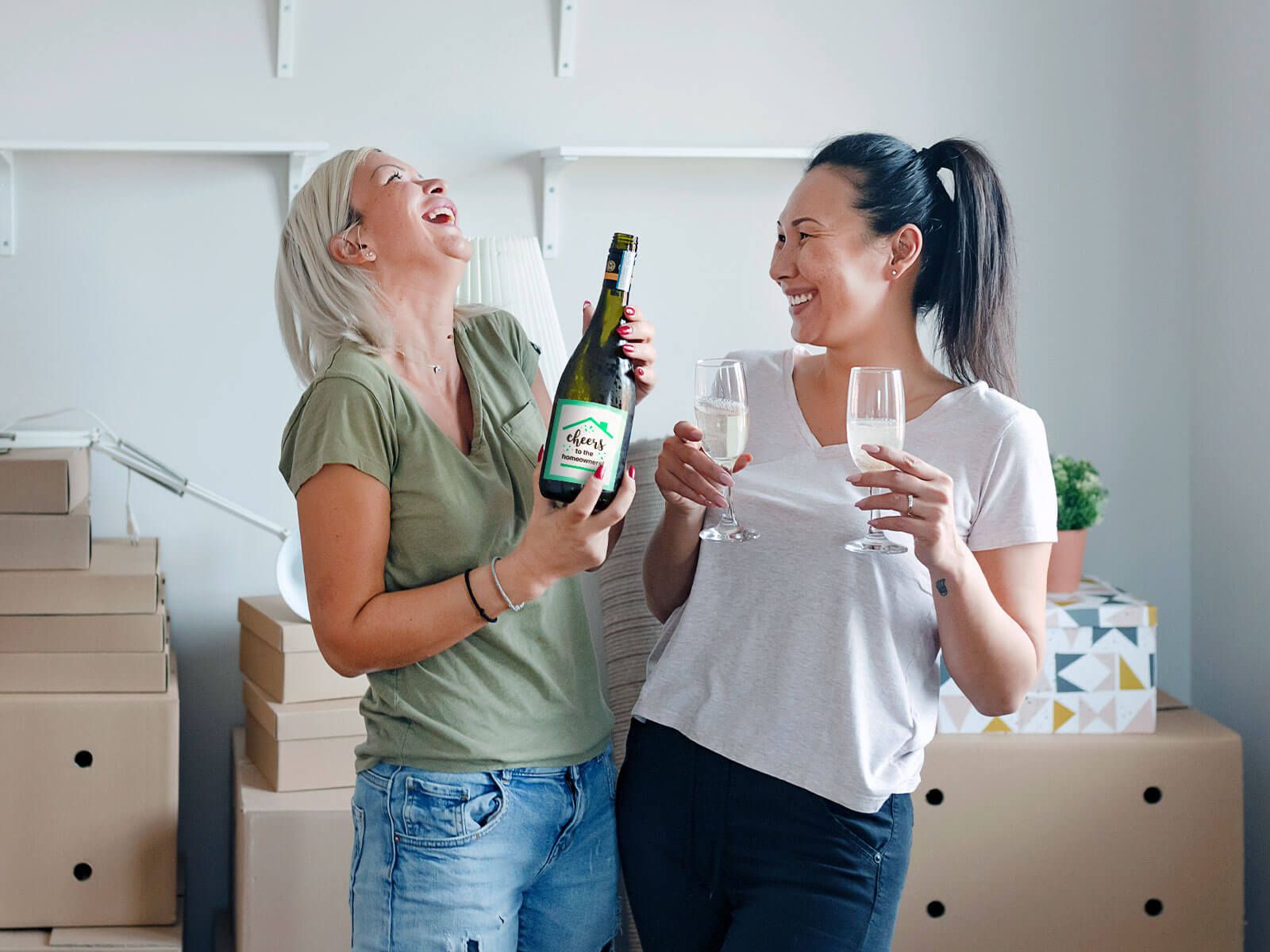 Two women holding a champagne bottle and glasses laughing with boxes in the background