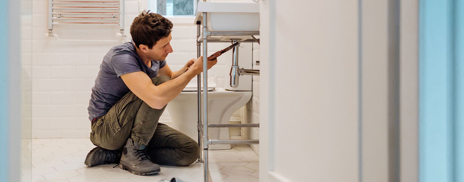 man kneeling at a sink, using a tool on the pipes