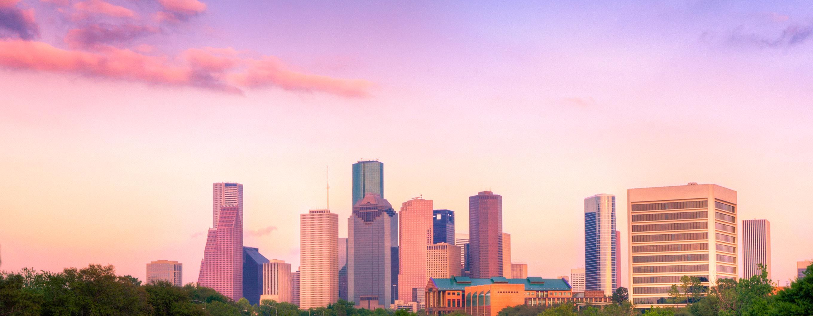 Image of the Houston city skyline with a purple and pink sky and trees in font of some of the buildings