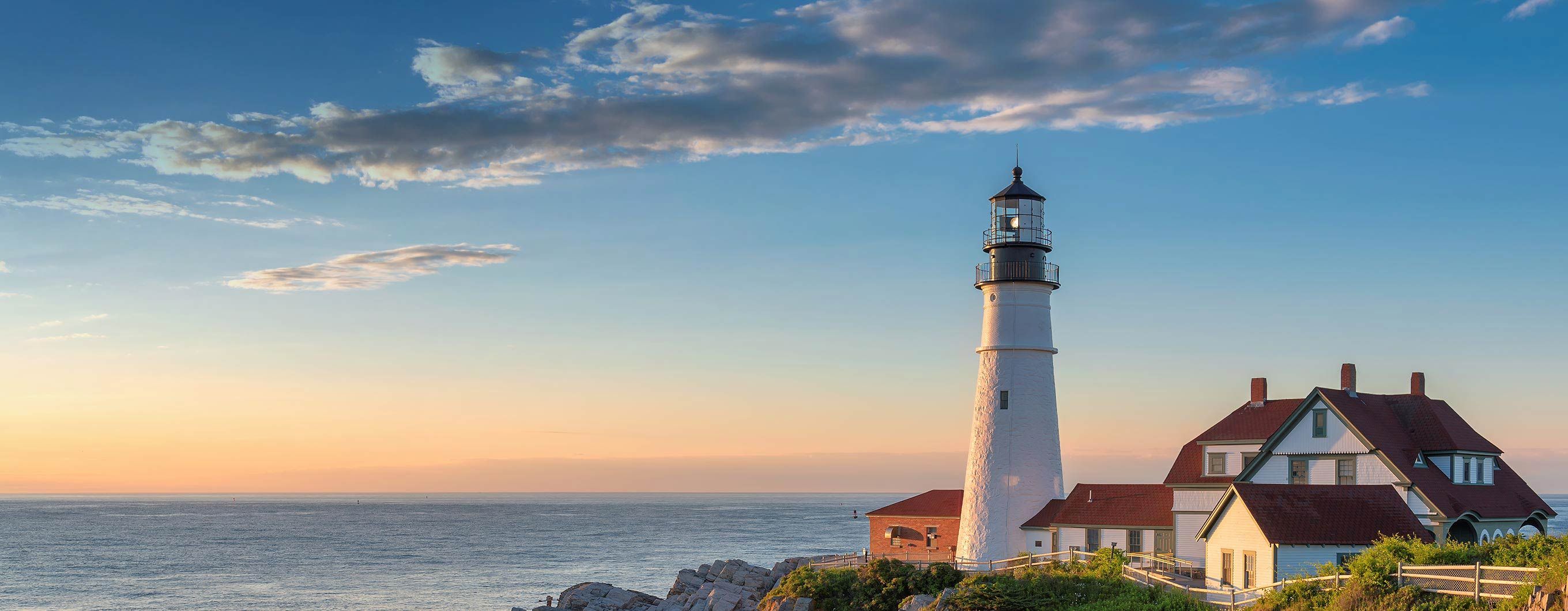 A white lighthouse sits on a rocky cliffside. It is sunset, and the sky is blue and yellow with a few clouds. 