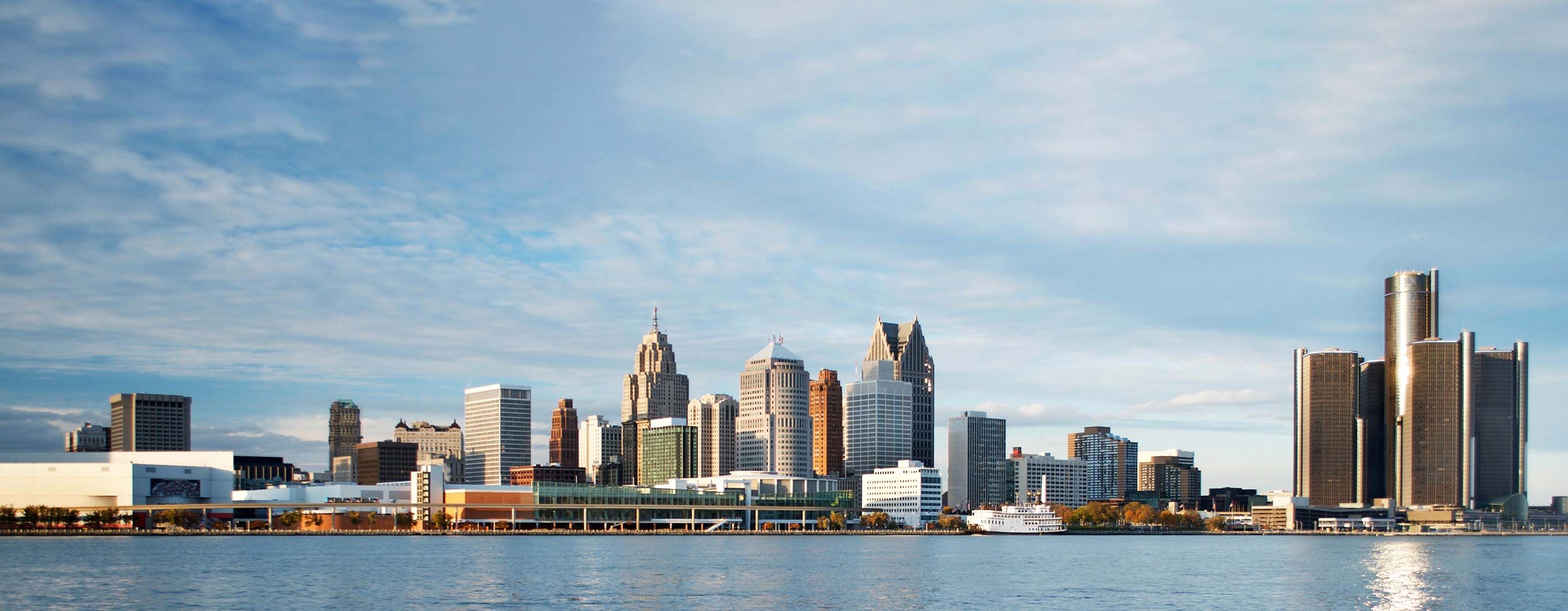 Image of the Detroit city skyline with a body of water in front and a blue sky with clouds behind