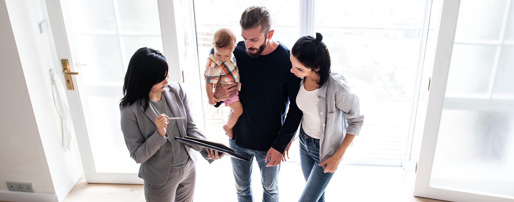 A woman and a man holding a baby talk to a woman in a suit holding a tablet