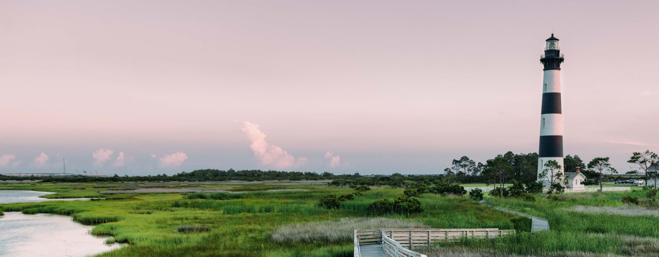 Image of a lighthouse and small building next to it surrounded by marshland, water and a bridge