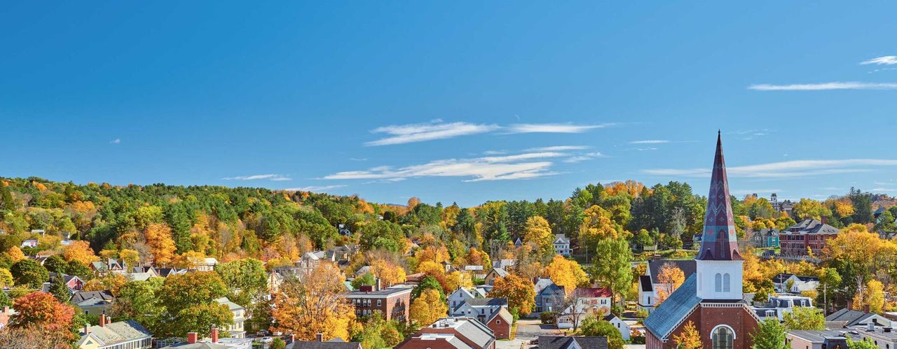 Image of a Vermont city showing the tops of homes and a church, with trees in the background in various fall colors
