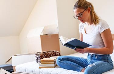 A woman sitting on a bed reading a book with boxes behind her