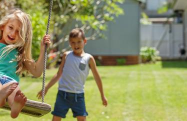 A young boy with brown hair pushes a young girl with blonde hair on a swing. Both are smiling. 