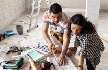 A man and a woman look over blueprint plans for a home. The man is measuring the plans with a tape measure. They are surrounded by tools. 