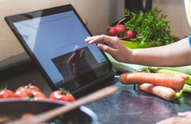 A person using a tablet in the kitchen with vegetables nearby