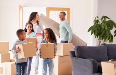 A family moves boxes into a home. A young boy and girl are in front smiling at each other, followed by another girl. The mom and dad are in the back of the room laughing. 