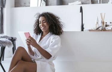 A woman sitting on the floor of her bathroom in a robe leaning on her tub while looking at her phone