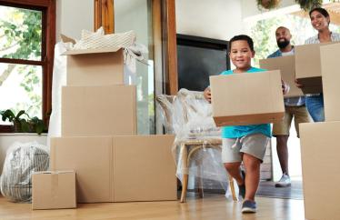 A little boy holding a moving box runs through a living room filled with moving boxes.