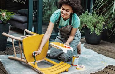 A woman paints a chair yellow on her wooden deck