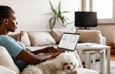 A Woman sits on a couch working on a laptop with a dog curled up by her side
