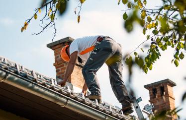 A man in construction gear stands on top of a house to fix the roof.