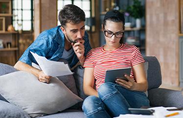 A man and woman sit on a couch with a tablet while reading about ways to save on their electric bill this summer.